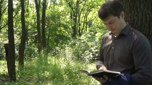 Νεαρός επιχειρηματίας διαβάζει ένα βιβλίο στο δάσος κοντά σε ένα δέντρο — Αρχείο Βίντεο