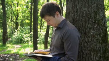 Genç işadamı ormanda bir ağacın yanında kitap okuyor.