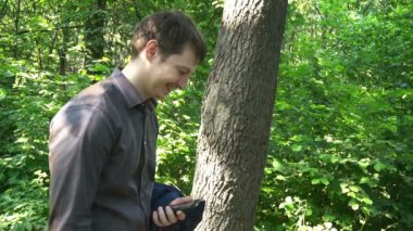 Genç adam ormanda duruyor, bir şeyler düşünüyor ve telefonda çalışıyor.