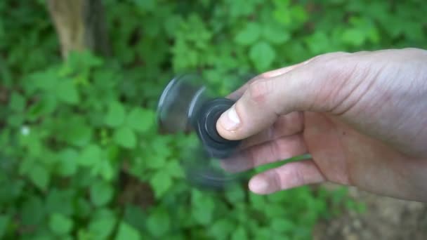 Black hand spinner, or fidgeting spinner, rotating on childs hand.grass background. Slow motion — Stock Video