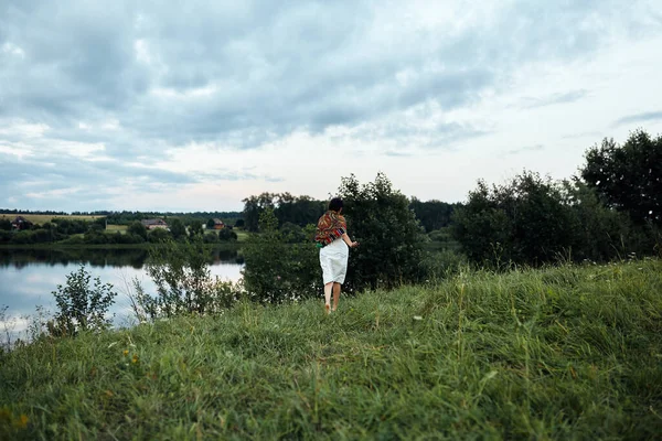 Una ragazza corre lungo un lussureggiante campo verde verso uno stagno — Foto Stock