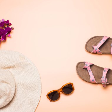 Hasır şapka, sandalet ve güneş gözlüğü pembe arka planda, kare şeklinde.