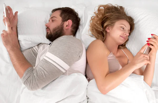 Молодая пара в постели смотрит телефон и игнорирует друг друга, лежа вместе — стоковое фото