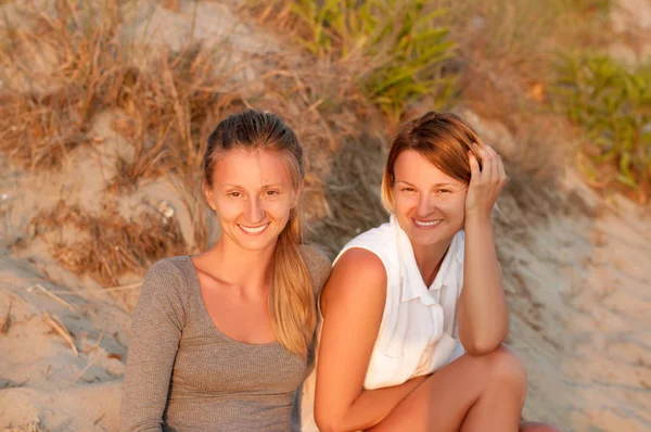 Gün batımında sahilde kum üzerinde oturan iki güzel kadın bak