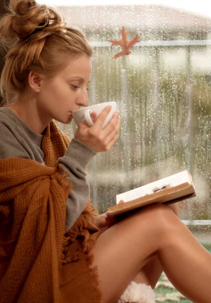 Sıcak çay içme ve sonbaharda bir kitap okuma sıcak bir ekose battaniye sarılı kadın