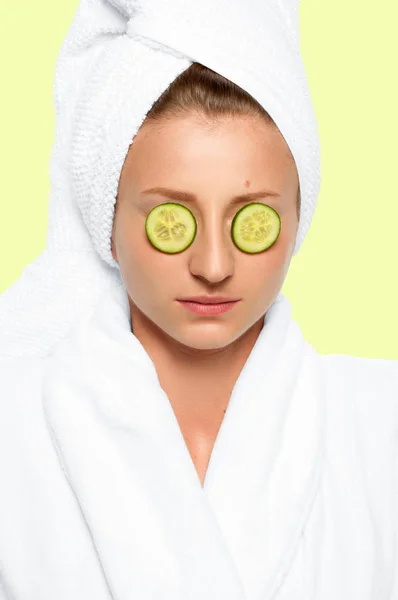 Beautiful woman with cucumbers on eyes and towel on her head on faded pastel background — Stock Photo, Image