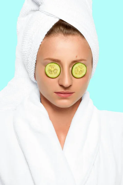 Beautiful woman with cucumbers on eyes and towel on her head on faded pastel background — Stock Photo, Image