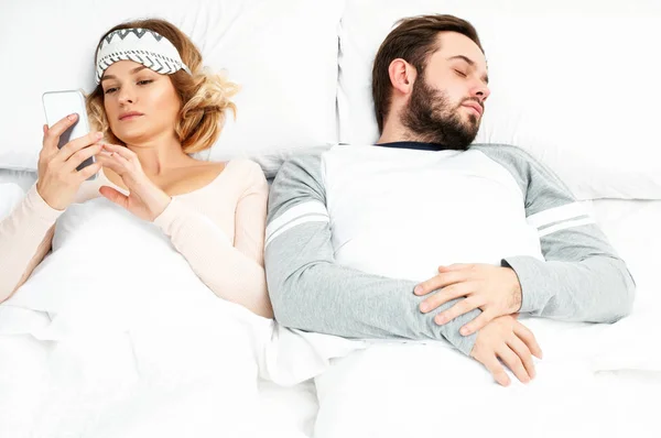 Mujer joven usando teléfono inteligente mientras su marido está durmiendo —  Fotos de Stock