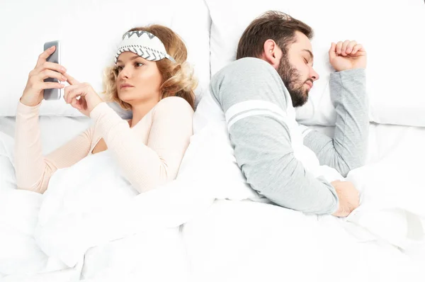 Νεαρή γυναίκα χρησιμοποιώντας smartphone, ενώ ο σύζυγός της στον ύπνο — Φωτογραφία Αρχείου