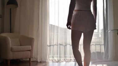 Genç kadın pencereye doğru hareket ediyor ve perdeleri açıyor. Evde dikilip, sabah şehir manzarasına bakıyor.