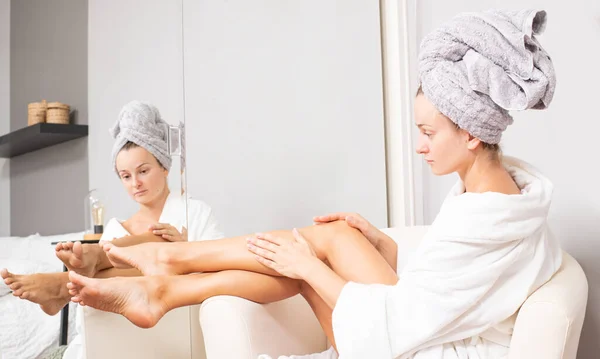 Beautiful girl in bathrobe is applying body lotion her legs. Depilation concept. — Stock Photo, Image