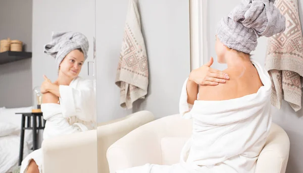 Beautiful woman in bathrobe after shower is applying body lotion on shoulder. — Stock Photo, Image