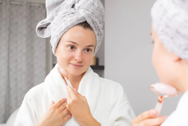 Beautiful woman is getting massage face using jade roller in front of the mirror at home — Stock Photo, Image