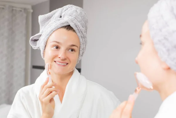 Beautiful woman is getting massage face using jade roller in front of the mirror at home — Stock Photo, Image