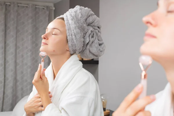 Красивая женщина получает массаж лицо с помощью нефритового ролика перед зеркалом в домашних условиях — стоковое фото