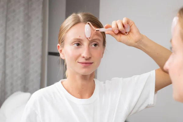 Krásná žena se dostává masáže obličeje pomocí nefritový válec před zrcadlem doma — Stock fotografie