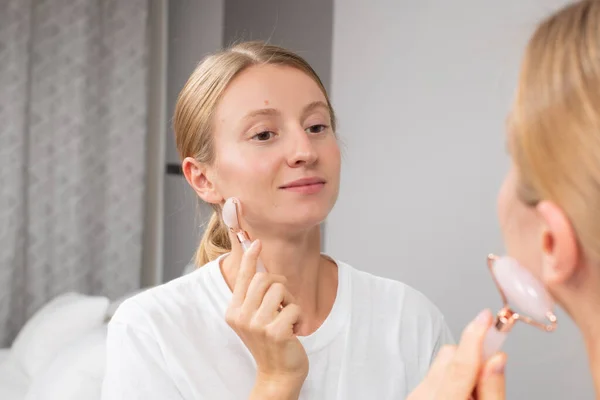 Mulher bonita está recebendo rosto de massagem usando rolo de jade na frente do espelho em casa — Fotografia de Stock