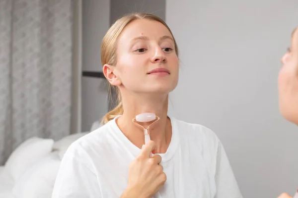 Beautiful woman is getting massage face using jade roller in front of the mirror at home — Stock Image Beautiful woman is getting massage face using jade roller in front of the mirror at home Stock Image