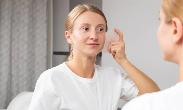 Beautiful woman is getting massage face using jade stone in front of the mirror at home — Stock Photo, Image