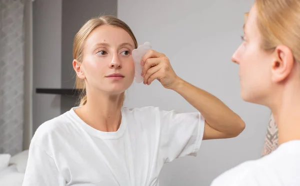 Mooie vrouw is het krijgen van massage gezicht met behulp van jade steen in de voorkant van de spiegel thuis — Stockfoto