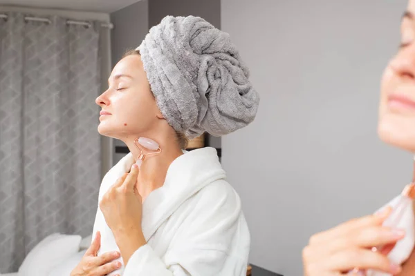 Beautiful woman is getting massage face using jade roller in front of the mirror at home — Stock Image Beautiful woman is getting massage face using jade roller in front of the mirror at home Stock Image