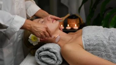 Kozmetik uzmanı, yeşim taşı kullanarak profesyonel yüz masajı yapıyor. Kadın güzellik merkezinde boyun masajı ve yüz masajı yaptırıyor..