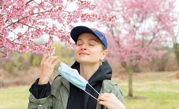 Beautiful woman takes off a medical mask and enjoying blooming tree after allergy