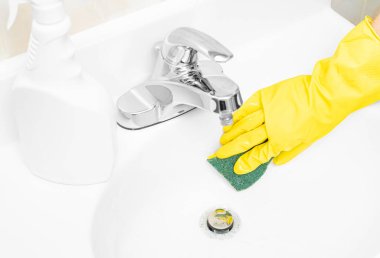 Sarı lastik eldivenli kadın banyo lavabosunu deterjan ve süngerle temizliyor.