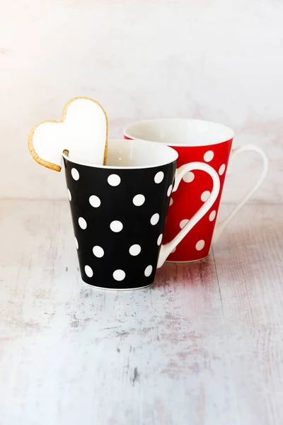 Ένα ζευγάρι μαύρες και κόκκινες polka διάστικτη φλιτζάνια καφέ, με μια καρδιά σχήμα σπιτικό μπισκότο στην άκρη πάνω από το λευκό φόντο ξύλου. — Φωτογραφία Αρχείου