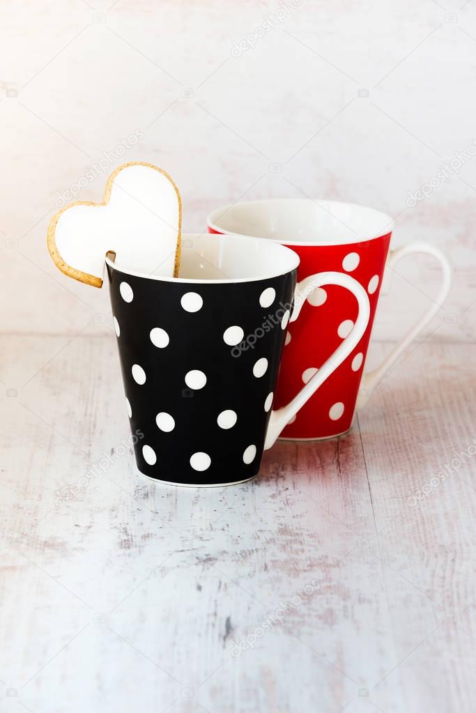 A pair of black and red  polka dotted coffee cups with a heart shaped homemade cookie on the edge over white wood background.