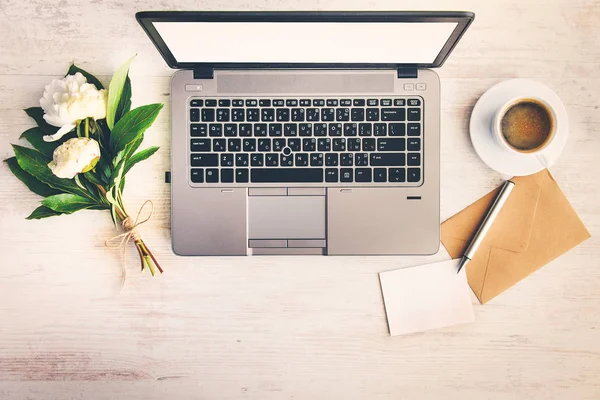 Draufsicht auf einen Schreibtisch mit Computer, Kaffeetasse, Umschlag, Blanko-Zettel und einem Strauß Pfingstrosen auf weißem Holzhintergrund. — Stockfoto