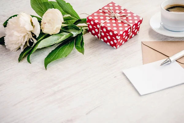 Coffret cadeau pointillé rouge, enveloppe kraft et carte de vœux vide, tasse à café et bouquet de fleurs de pivoine sur fond de bois blanc. Espace de copie . — Photo