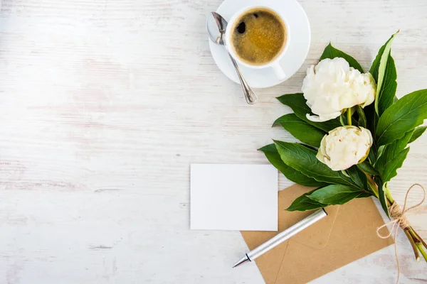 Vue du dessus d'une carte de vœux vide, d'une enveloppe kraft, d'un bouquet de pivoines et d'une tasse de café sur fond rustique en bois blanc . — Photo