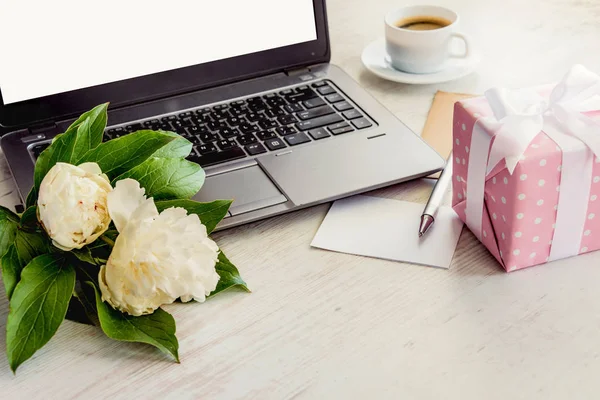 Vue latérale d'un pont avec ordinateur, bouquet de fleurs de pivoines, tasse de café, carte vide et boîte cadeau en pointillés roses. Fond en bois rustique blanc. Perspective romantique et féminine . — Photo