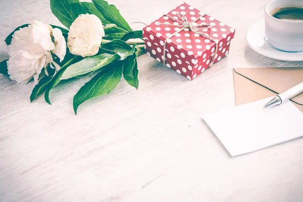 Coffret cadeau pointillé rouge, enveloppe kraft et carte de vœux vide, tasse à café et bouquet de fleurs de pivoine sur fond de bois blanc. Espace de copie. Effet vintage . — Photo