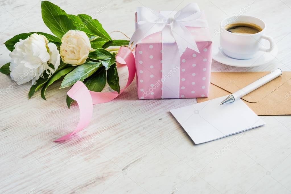 Pink dotted gift box, empty greeting card, kraft envelope, peonies bouquet and coffee cup over white wooden rustic table. Romantic holiday and event concept.