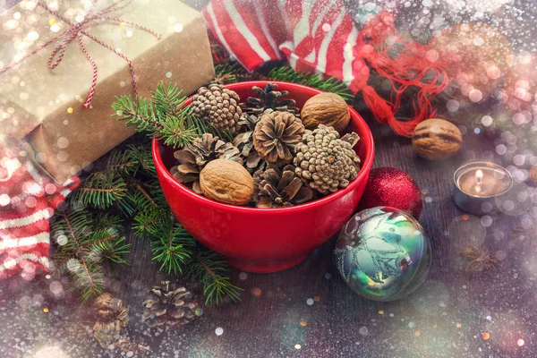 Dekorację świąteczną - czerwony miskę pełną szyszek jodły, pudełko zawinięte w papier pakowy, gałęzie sosnowe, Świeca, orzechy, anyż, jabłka, Boże Narodzenie zabawki i szalik biało -czerwone paski zima. Boże Narodzenie Motyw. — Zdjęcie stockowe