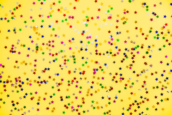 Scattered star shaped colorful glittering confetti over yellow background. — Stock Photo, Image