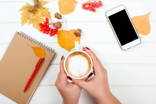 Mãos femininas segurando uma xícara de cappuccino. Fundo de outono, notebook com caneta vermelha, telefone móvel na mesa de madeira . — Fotografia de Stock