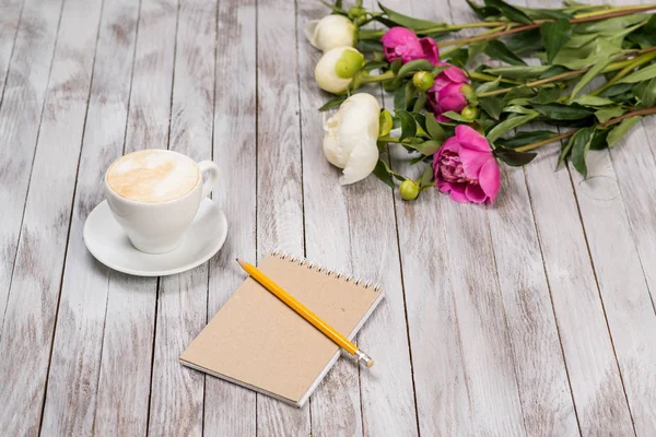Notebook z ołówka obok kawy i piwonie kwiaty na podłoże drewniane. — Zdjęcie stockowe