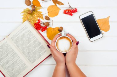 Bir fincan kapuçino tutan eller. Sonbahar arka plan, kırmızı kalem, ahşap masa üstünde hareket eden telefon ile defter.