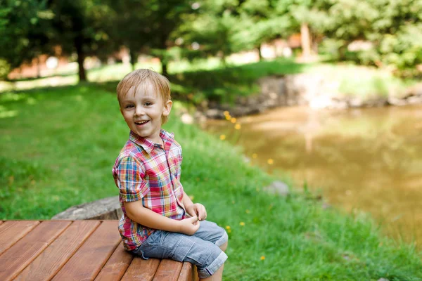 호수 근처 벤치에 앉아 웃는 행복 한 소년. 여름 시간 주말 분위기. — 스톡 사진