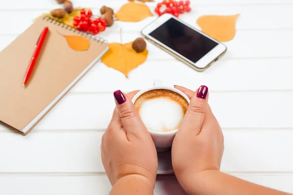 Mãos femininas segurando uma xícara de cappuccino. Fundo de outono, notebook com caneta vermelha, telefone móvel na mesa de madeira . — Fotografia de Stock
