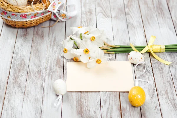 Ovos de Páscoa em cesta, cartão de saudação vazio, buquê de narcisos no fundo de madeira. Decorações de Páscoa . — Fotografia de Stock