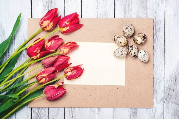 Ovos de Páscoa, belas tulipas vermelhas, nota em branco no fundo de madeira . — Fotografia de Stock