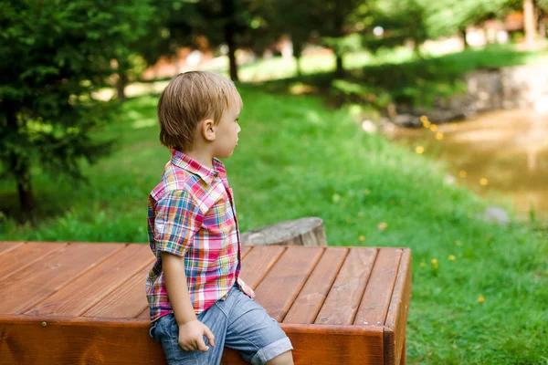 Lachende gelukkige jongen zitten op bankje in de buurt van lake. Zomertijd weekend stemming. — Stockfoto