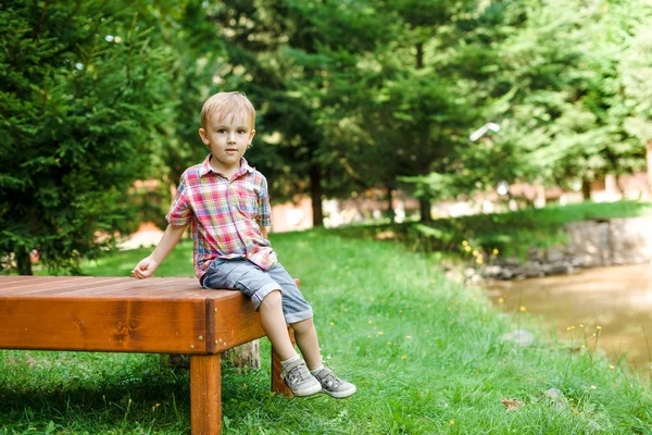 Sonriente niño feliz sentado en el banco cerca del lago. Tiempo de verano humor fin de semana . — Foto de Stock