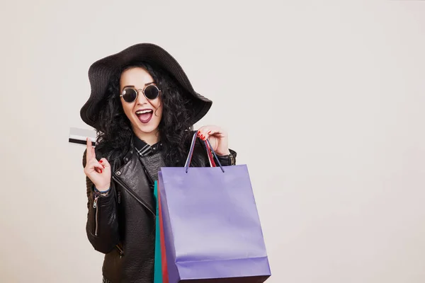 Mujer joven de moda en sombrero negro con tarjeta de crédito y bolsas de compras de colores sobre fondo de estudio blanco . — Foto de Stock