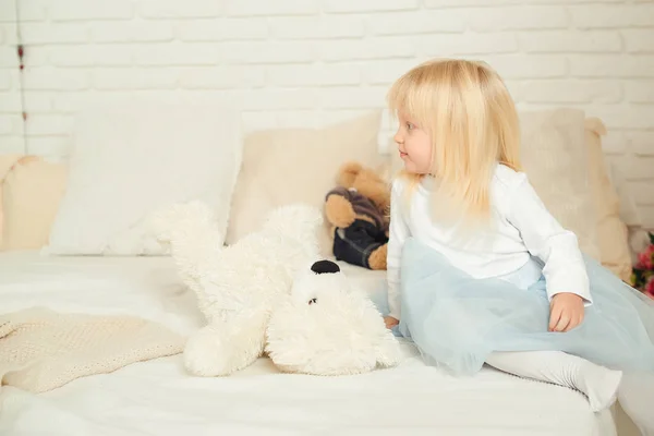 Cute toddler girl sitting on the bed with her soft toys in a light room. Happy birthday concept. — Stock Photo, Image