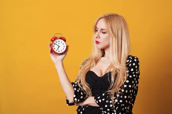 Pensive blondy woman looking at alarm clock over yellow background.